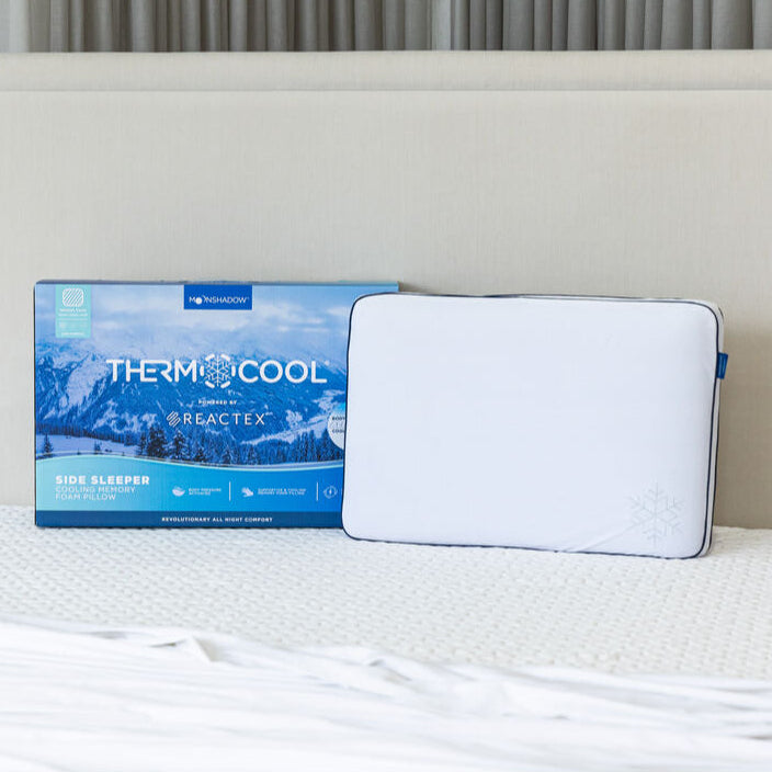A white pillow designed for side sleepers, with a cooling feature, placed on a bed with a headboard. The packaging is visible and displays the product name 'Thermocool'.